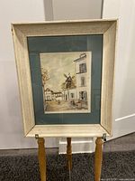 Framed vintage lithograph depicting an urban scene with buildings and windmill, with white distressed wooden frame