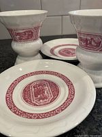 Close-up photo of two white porcelain Rüdesheimer cups and matching saucers, featuring red scenic decals depicting traditional German scenes.
