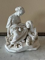 Front view of ceramic sculpture showing woman holding broken shoe and child sitting at her side on base with floral details.