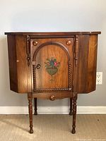 Front view of wooden sewing table showing hand painted floral design on central door, storage compartments on sides, turned legs, and some surface wear.