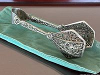Pair of large silver plated serving tongs with detailed floral and ornamental patterns, placed on a turquoise cloth.
