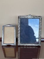 Two antique sterling silver picture frames side by side on a wooden surface against beige wall. Larger frame shows glass with tarnished reflective surface, smaller frame has no glass.