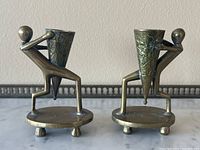 Pair of brass figural candle holders showing full front view with human figures holding cone-shaped cups decorated with embossed designs, placed on oval bases with four feet.