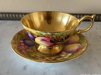 Angled side view of the gold interior cup and matching saucer showing hand-painted fruits and gold rim.