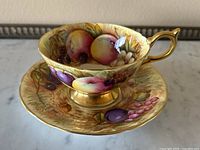 Overall side view of the teacup on the saucer showing detailed hand-painted fruit and gold accents.