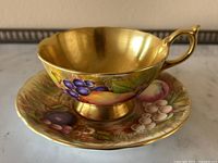 Front-side view of teacup and saucer showing hand-painted fruit design and gold interior of cup