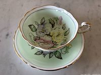Overhead view of teacup nestled in saucer showing mint green background with purple, pink, and yellow pansy floral design and gold trim around edges.