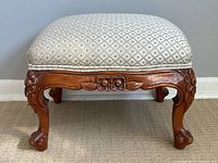 Front view of ornately carved antique wooden footstool showing detailed floral carvings on apron and curved legs ending in claw feet, topped with patterned upholstered cushion.