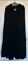 Full frontal view of the vintage black velvet opera cape hanging on a door, showing its length and drape.