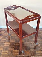 Full view of the mahogany bar cart angled to show the carved top tray with glass surface and lower shelf with cracking.