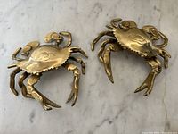 Two solid brass crab shaped ashtrays placed side by side on a marble surface, showing entire crab shape and size.