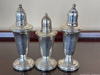 Lot of three sterling silver salt and pepper shakers in silver tone with varied lid designs, shown together on wood surface.