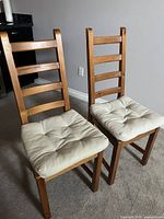 Two wooden kitchen chairs side by side showing overall appearance, each with a beige cushioned seat pad.