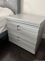 Nightstand in place next to bed showing two drawers with finger-pull design and grey painted finish.