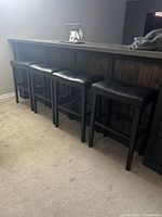 Four black bar stools with padded seats positioned against a bar counter on carpeted floor.