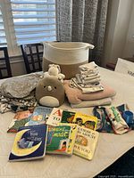 Photo of the lot showing the basket, plush toys, folded clothes and blankets, and board books spread on a surface.