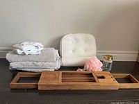 Overview of the bath items: stacked light grey towels, white cushioned bath pillow, pink bath sponge, small jar with gold lid, and wooden bath tray on floor.