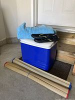 Photo showing blue Igloo cooler with white lid, large blue towel on top, men's black water shoes with neon green detailing, two rolled straw beach mats on the floor beside cooler.
