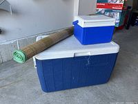 Photo showing two blue Coleman coolers stacked, large cooler at bottom and small cooler on top, with a rolled green straw mat beside them.