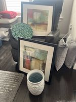 Two matching framed abstract prints seen resting on a black chair, prints sized 22x22 inches, one positioned behind the other, with a white ceramic vase and blue mosaic decorative plate in front.