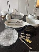 Overview of assorted serving bowls, trays, cutlery, sugar jar, cheese knives and butter pats on black countertop.