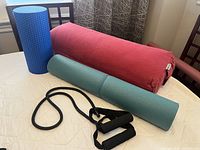 Blue foam roller, red yoga bolster, green rolled yoga mat, and black tension/exercise band shown on a bed.