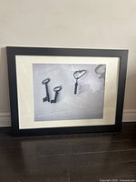 Framed photo print showing two vintage keys and one key-shaped object casting shadows on a light background in a black frame with white matting, leaning against a wall above a wooden floor.