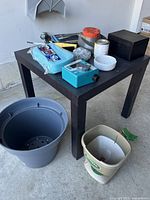 Small black square table with various gardening accessories on top including a fertilizer spreader, blue water timer, black Faraday box for keys, white bowl, and a gray plastic planter pot and beige spreader container on the floor next to table.