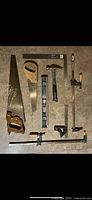 Photo of laid out hand tools including two saws, hammer, level, bar clamps, try square, and ruler, showing the full set of visible tools.