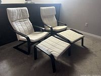 Two IKEA Poäng lounge chairs with matching footstools set on carpet, showing black bentwood frames and white cushions with headrests.