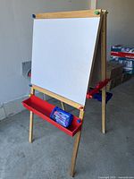 Front side of the wooden kids easel showing whiteboard and red tray containing blue pencil case with markers.