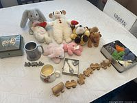 Photo showing the entire lot arranged on a white surface including stuffed animals, toy train, pewter items, ceramic mugs, and jewelry box.