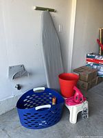 Photo showing blue laundry basket with small items inside, pink handheld steamer on small white stool, tall red plastic container, and full-sized ironing board with grey cover against wall.