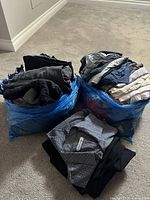 Two large blue bags filled with assorted men's clothing items such as jeans, t-shirts, pants, and dress shirts, along with a grey patterned dress shirt and dark pants placed on carpet.