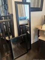 Two black framed mirrors, one tall rectangular and one smaller square, leaning against a wall beside furniture.