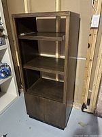 Tall brown oak veneer AV cabinet with open shelves, a pull-out shelf near the top, and closed lower cabinet with doors. Positioned in basement with unfinished walls.