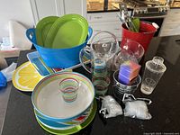 Blue punch bowl, colorful bowls and plates, plastic straws, clear pitchers, and drinking cups arranged on a countertop.