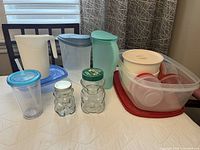 Several plastic containers and pitchers displayed on a table, including two vintage glass peanut butter bear jars with lids, plastic pitchers in white and light green, a clear plastic cup with blue lid and straw, and multiple plastic containers with red and white lids.