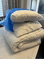 Stack of three white pillows on top of folded white duvet insert with blue plastic on top pillow.