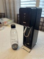 Photo of SodaStream carbonation machine with reusable transparent plastic bottle with black cap attached to front.