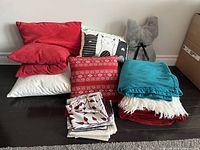 Full group view of 8 throw pillows in various colors and patterns, and 2 folded throw blankets in teal and red with white fringe on a dark floor.