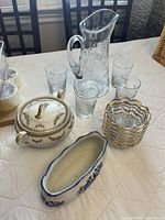 Photo showing clear etched glass pitcher with handle, six matching clear etched drinking glasses, stack of scalloped edge glass bowls with gold accents, decorative lidded ceramic container with gold detailing and a blue and white ceramic dish on a table.