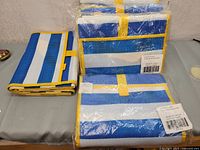 Five blue and white striped beach mats with yellow edging, one unwrapped showing texture and the rest stacked or folded and packaged.