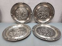 Four German pewter wall hanging plates arranged side by side, showing detailed seasonal scenes in relief.