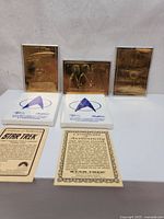 Three 22ct gold-plated Star Trek collectible cards displayed upright with two certificates of authenticity and two plastic stands with Star Trek logo.