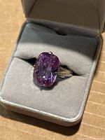 Top view of the ring inside a grey velvet box showing the large purple gemstone and four-prong setting.
