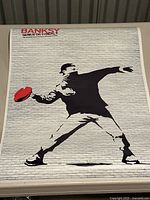 Photo of a Banksy print showing a masked man throwing a red object with text 'BANKSY GENIUS OR VANDAL' on white brick wall background