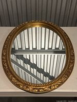 Photo of round mirror with ornate gold frame on a white table against corrugated metal background, showing mirror surface and frame detail.