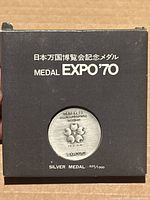 Black display box with Japanese text, EXPO 70 silver medal label, and circular cutout revealing the silver medal inside with Expo 70 emblem and inscriptions.