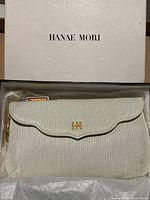 White textured purse with scalloped flap, gold 'HM' logo, and gold chain strap inside Hanae Mori box with tissue paper.